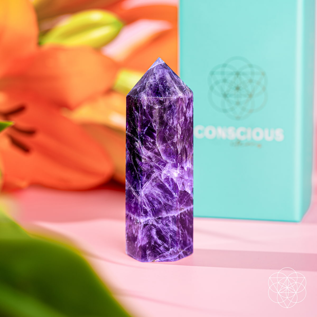 The Cycle Breaker - Chevron Amethyst Point of Transformation by Conscious Items stands upright on a pink surface for stress relief, with orange lilies and green leaves in the background, next to a blue Conscious Items box.