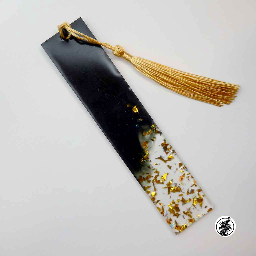 A rectangular black resin bookmark with a gold tassel, featuring a gradient design that transitions from solid black to a white section filled with scattered gold foil flakes, on a light gray background.