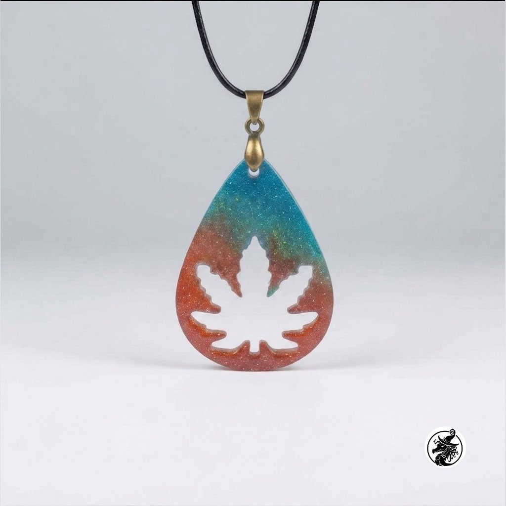 A teardrop-shaped pendant necklace with a black cord, featuring a blue-to-orange gradient background with a white maple leaf silhouette in the center and a gold-toned bail.