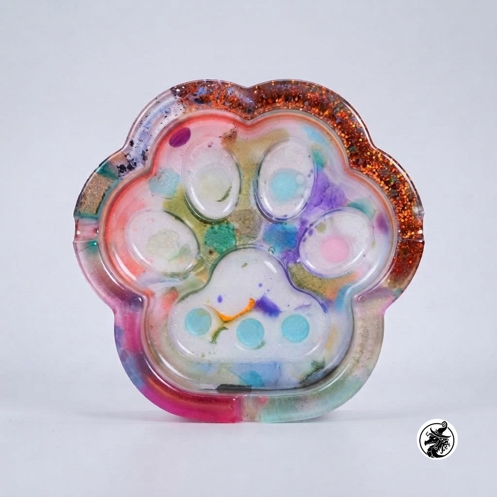 A paw-shaped resin tray with a glittery copper-orange rim and a multicolored marbled interior featuring a paw print design, photographed on a light gray background.