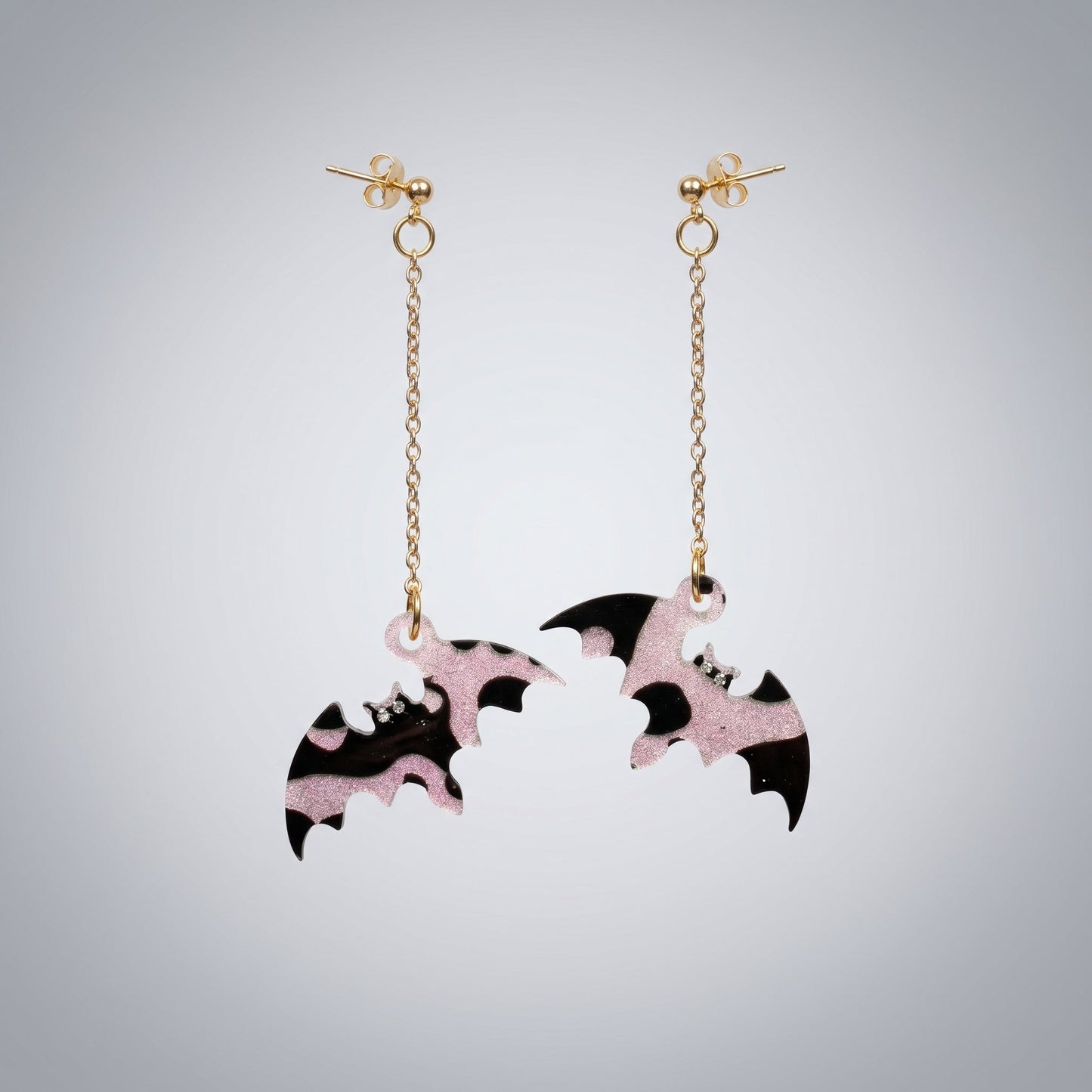 Dangle earrings featuring black and pink glitter resin bats with gold chains on a dark brushed metal surface.