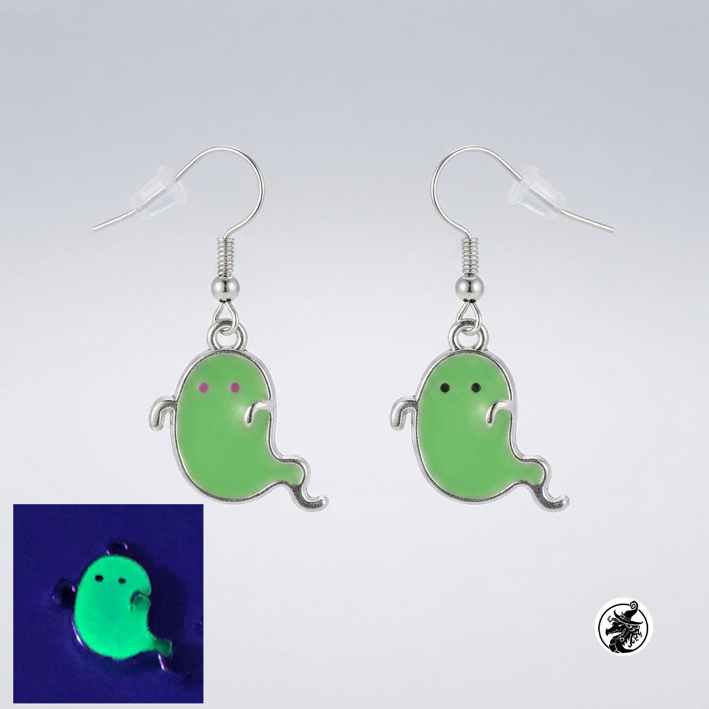 A pair of dangling ghost-shaped earrings with silver hooks and light green enamel, shown on a light gray background. A small inset at lower left shows the earring glowing bright green in the dark, and a small logo appears in the lower right corner.