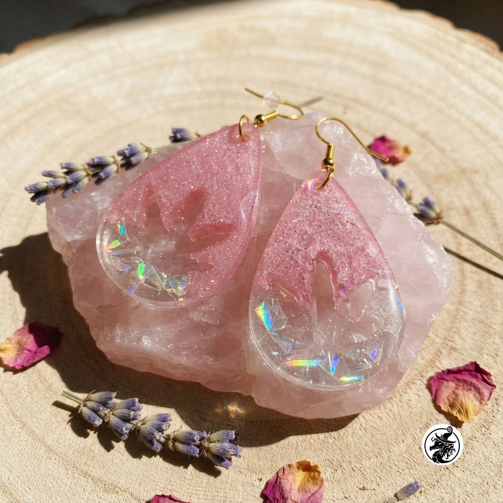 A pair of teardrop-shaped pink resin earrings with a cannabis leaf cutout and holographic glitter, displayed on a pink crystal cluster with dried lavender and rose petals on a wood slice background.