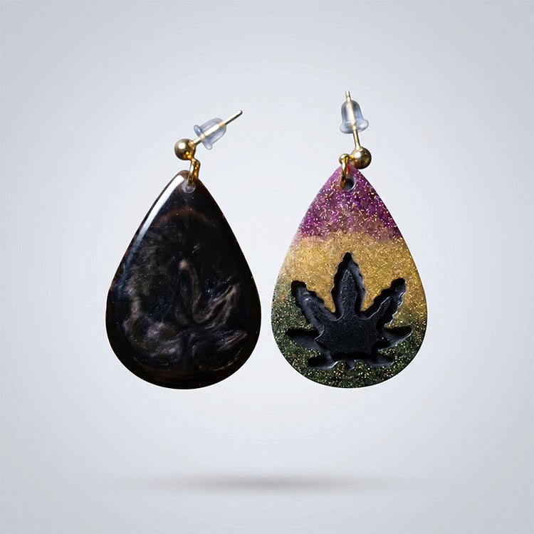 Vertical view of two resin earrings: one with a tri-color glitter leaf motif and one with a dark smoke-like swirl.