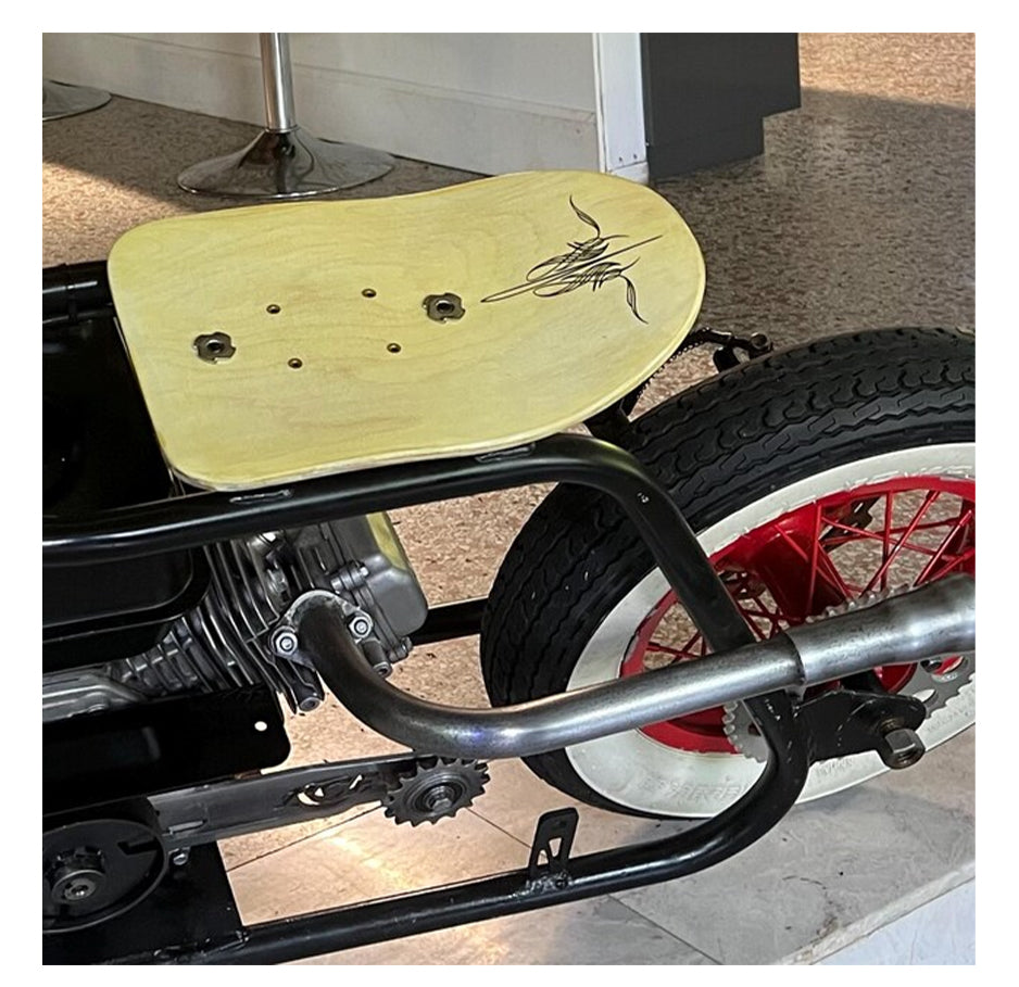 A close-up of the Pinstripe Cox - Style 01 by GorgeousDragon Designs LLC., a custom motorcycle seat with a black artistic design on wood, mounted on a metal frame near a rear tire with red spokes and white sidewalls.