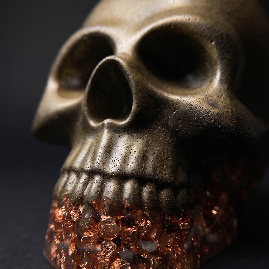 The Brass Infused Distressed Skull Paperweight by GorgeousDragon Designs LLC. features orange crystal-like stones in the jaw, making it a bold gothic decor or desk accessory—all set against a sleek black backdrop.