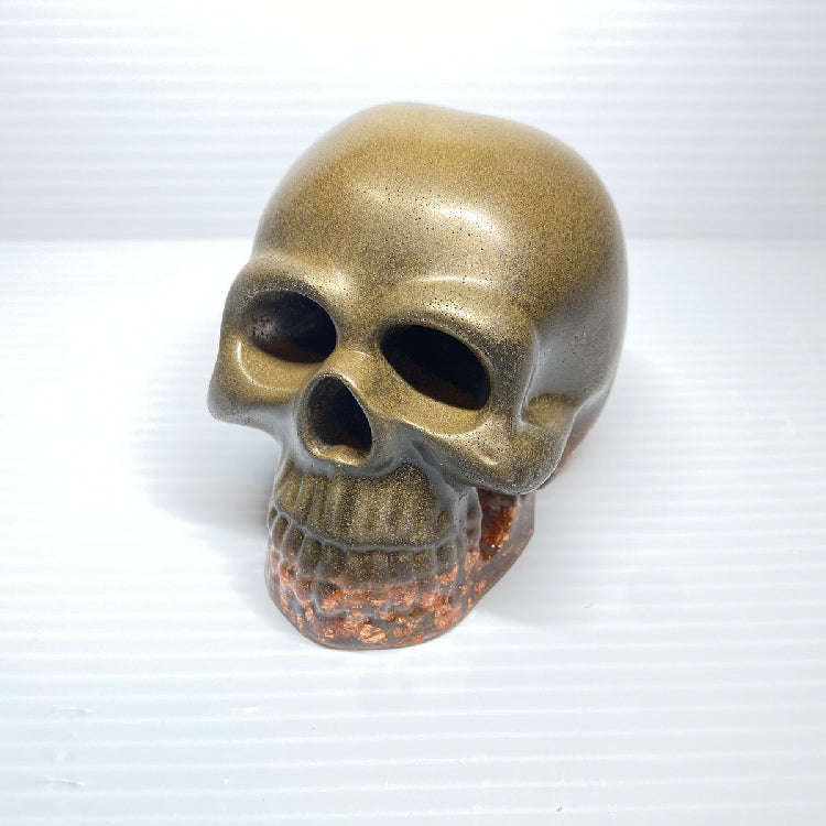 The Brass Infused Distressed Skull Paperweight by GorgeousDragon Designs LLC features a metallic gold finish fading into reddish-brown, set against a white textured background—ideal as gothic decor or a unique office desk accent.