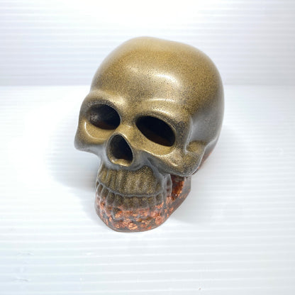 The Brass Infused Distressed Skull Paperweight by GorgeousDragon Designs LLC features a metallic gold finish fading into reddish-brown, set against a white textured background—ideal as gothic decor or a unique office desk accent.
