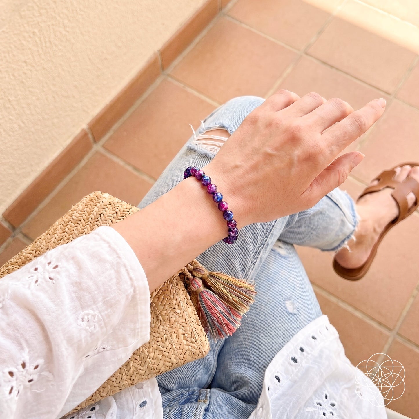 A person in a white eyelet blouse and ripped jeans sits on tiled floor, holding a woven tassel clutch. They wear brown sandals and the Destiny Activator - All-Galaxy Tiger’s Eye Bracelet by Conscious Items, symbolizing cosmic destiny.