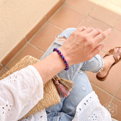 A person in a white eyelet blouse and ripped jeans sits on tiled floor, holding a woven tassel clutch. They wear brown sandals and the Destiny Activator - All-Galaxy Tiger’s Eye Bracelet by Conscious Items, symbolizing cosmic destiny.