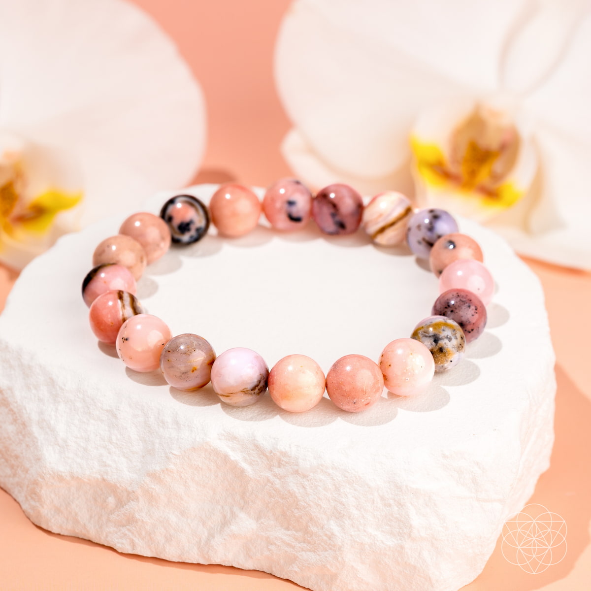 The Repair My Heart - The Peruvian Pink Opal Bracelet by Conscious Items, featuring round pink and purple marbled stones, is showcased on a white stone with two white flowers and a peach backdrop, symbolizing heart healing.
