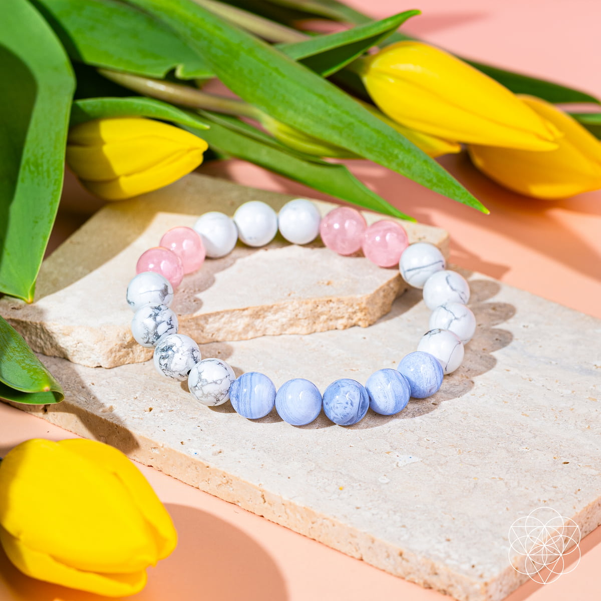 The Conscious Items 3AM Thoughts Off - Deep Sleep Trio Bracelet, featuring white, light blue, and pink calming stones, is displayed on a stone slab surrounded by yellow tulips atop a pale pink surface.