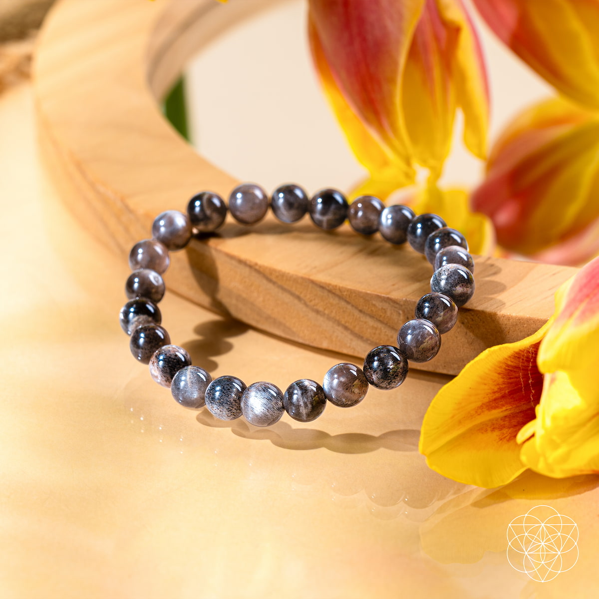 The Empath’s Cord Cutter – Black Moonstone Bracelet by Conscious Items, featuring dark, shiny beads, sits on light wood with yellow and red tulip petals—symbolizing new beginnings. A geometric white logo is shown in the lower right corner.