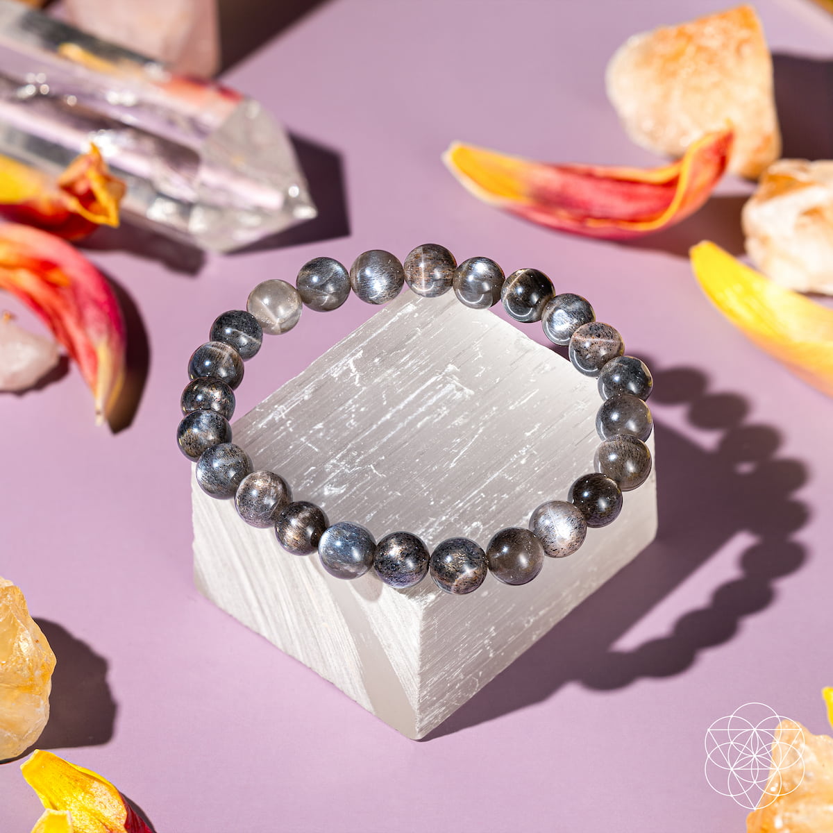 The Empath’s Cord Cutter – Black Moonstone Bracelet by Conscious Items rests on a white crystal block, surrounded by vibrant crystals and colorful petals, evoking new beginnings on a pastel purple surface.