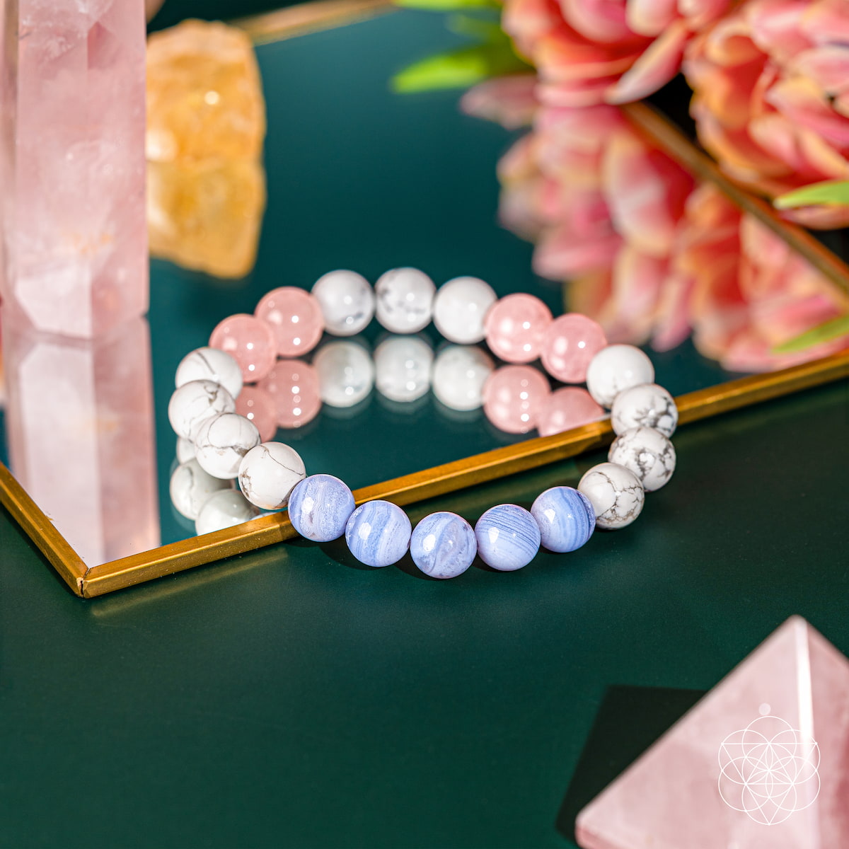 The 3AM Thoughts Off - Deep Sleep Trio Bracelet by Conscious Items, featuring white, pink, and light blue calming stones for sleep, rests on a mirrored surface with reflections of pink crystals and flowers.