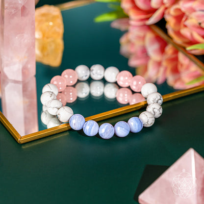 The 3AM Thoughts Off - Deep Sleep Trio Bracelet by Conscious Items, featuring white, pink, and light blue calming stones for sleep, rests on a mirrored surface with reflections of pink crystals and flowers.