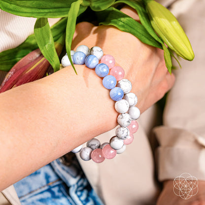 A close-up of a wrist wearing Conscious Items' 3AM Thoughts Off - Deep Sleep Trio Bracelet—three beaded bracelets in white, pink, and light blue—holding green leaves and a flower bud. The person is dressed casually in light colors.