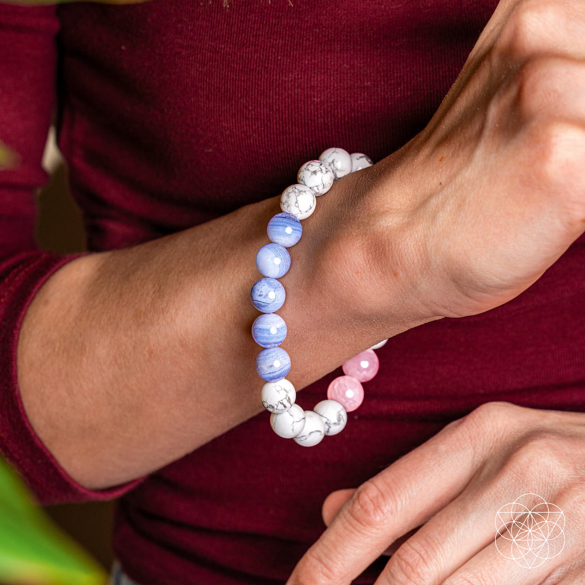 A person wearing a maroon long-sleeve top showcases the Conscious Items 3AM Thoughts Off - Deep Sleep Trio Bracelet with calming white, blue, and pink marbled beads on their wrist. A geometric logo appears in the bottom right corner.
