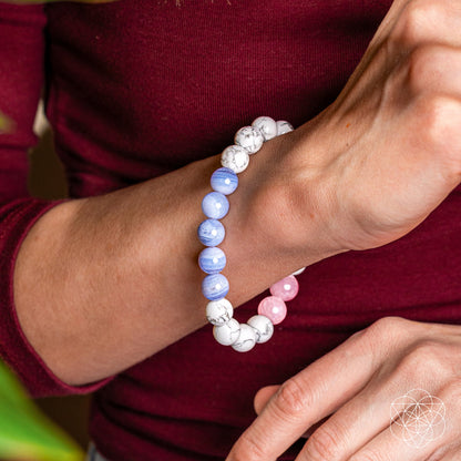A person wearing a maroon long-sleeve top showcases the Conscious Items 3AM Thoughts Off - Deep Sleep Trio Bracelet with calming white, blue, and pink marbled beads on their wrist. A geometric logo appears in the bottom right corner.