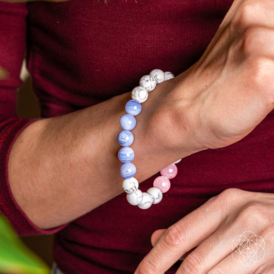 A person wearing a maroon long-sleeve top showcases the Conscious Items 3AM Thoughts Off - Deep Sleep Trio Bracelet with calming white, blue, and pink marbled beads on their wrist. A geometric logo appears in the bottom right corner.