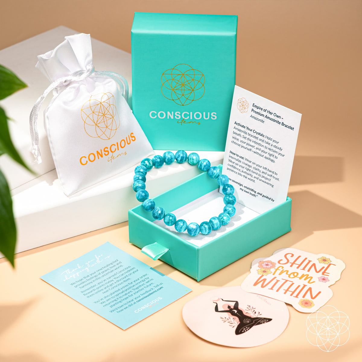 The Empire of Her Own – Premium Blue Amazonite Bracelet by Conscious Items rests in an open teal box, surrounded by a white pouch, informational cards, and stickers with uplifting messages on a light beige surface.