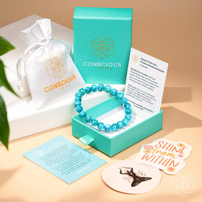 The Empire of Her Own – Premium Blue Amazonite Bracelet by Conscious Items rests in an open teal box, surrounded by a white pouch, informational cards, and stickers with uplifting messages on a light beige surface.
