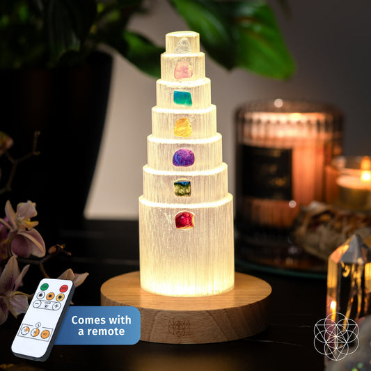 The Stress Shield Lamp - 7 Stones of Healing by Conscious Items features a glowing selenite tower with seven gemstones on a wooden base, remote control included. Soft lighting and plants complement this calming decor piece.