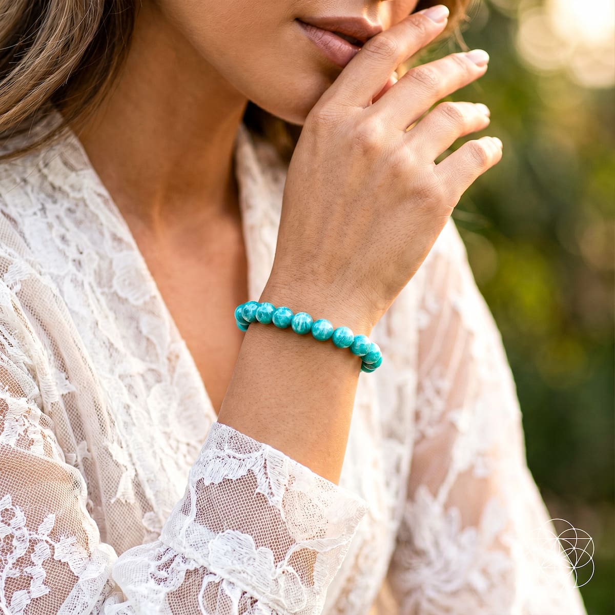 A woman in a white lace top touches her face, displaying the Empire of Her Own – Premium Blue Amazonite Bracelet by Conscious Items on her wrist, set against a blurred outdoor backdrop.