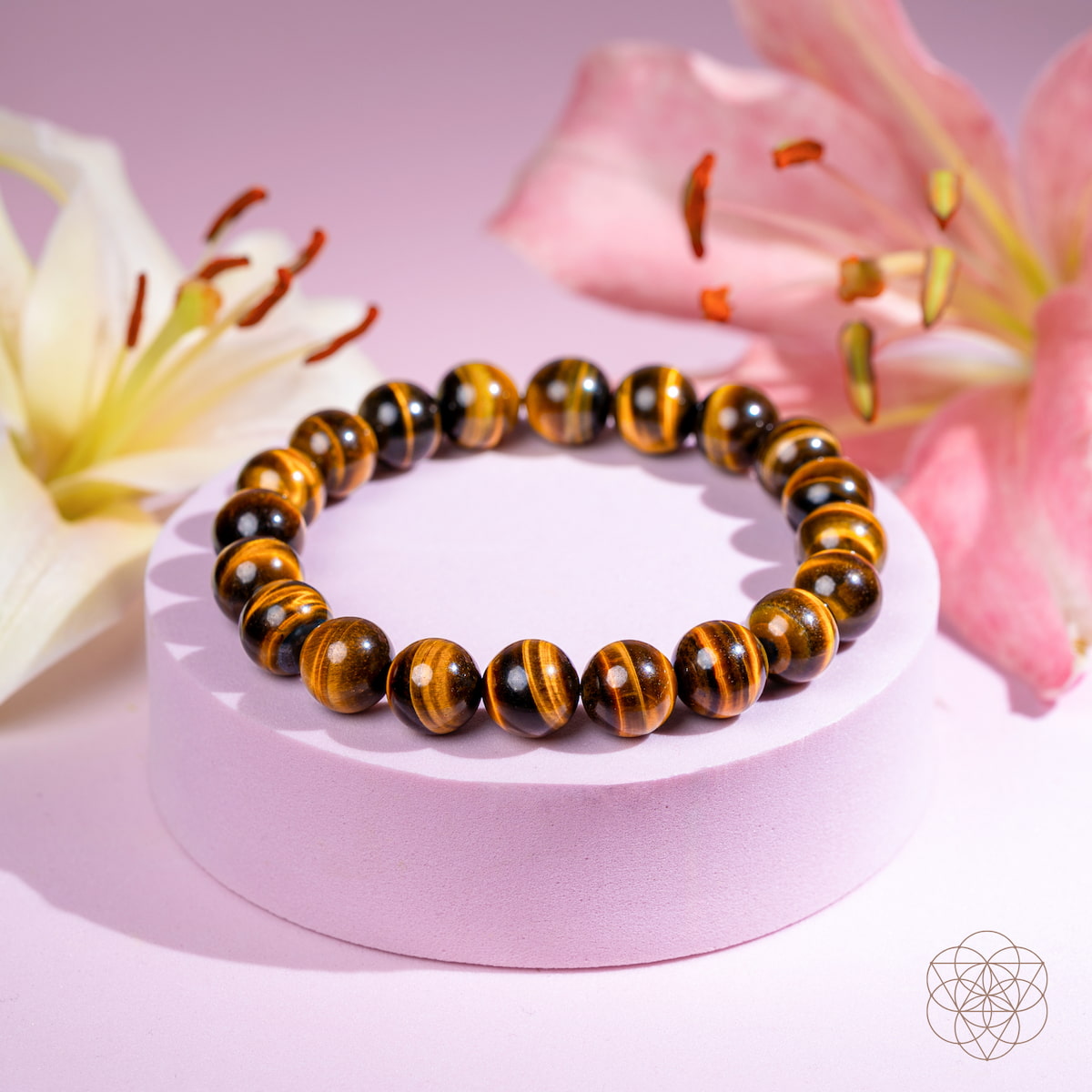 The Built to Win – 7A Grade Tiger’s Eye Bracelet by Conscious Items is displayed on a round, pale pink stand with white and pink lilies in the background, set against a soft pink backdrop.