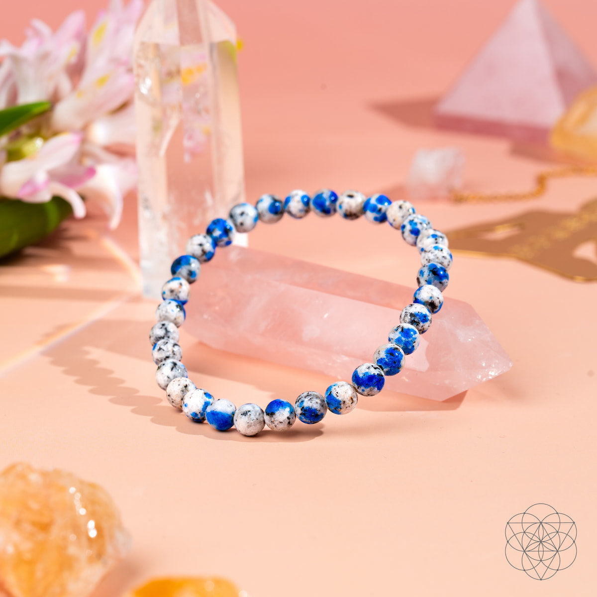 The Rare K2 Stone Bracelet of Spiritual Adventure | 6mm by Conscious Items, with blue and white beads, is shown on a pink surface beside crystals, flowers, and pastel geometric decor.