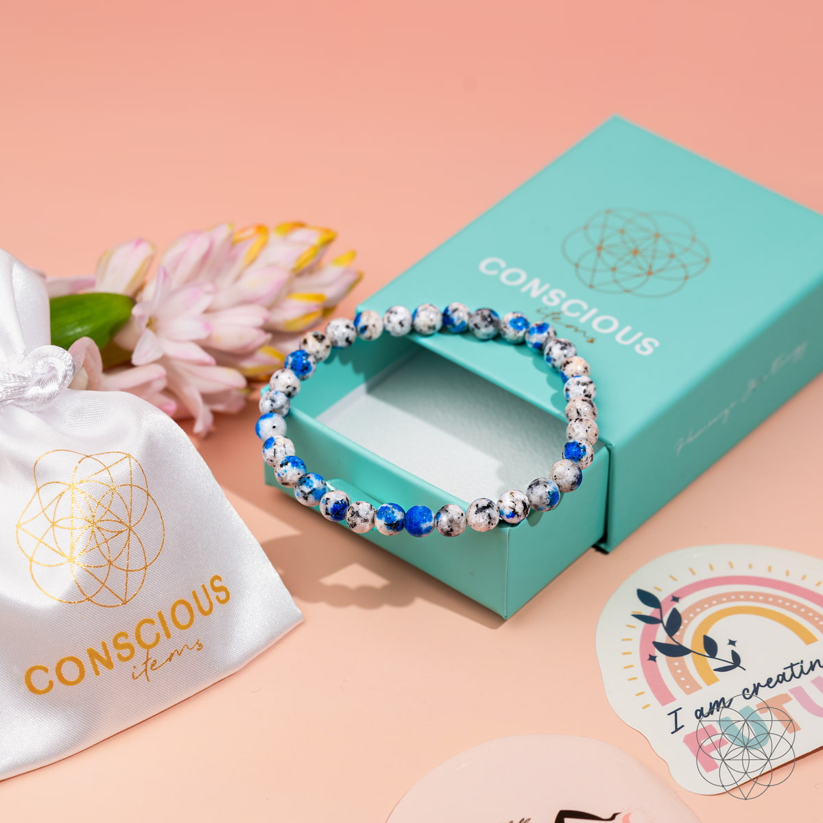 The Rare K2 Stone Bracelet of Spiritual Adventure | 6mm by Conscious Items rests on a turquoise Conscious Items box, near a white satin pouch, a pink flower, and colorful stickers on a peach background.