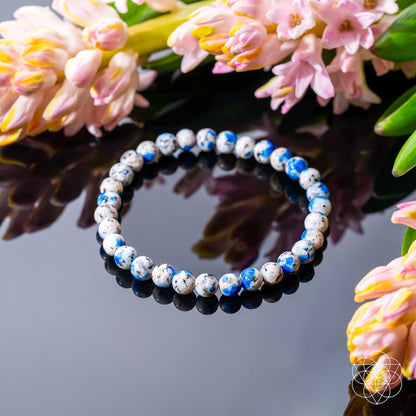 The Rare K2 Stone Bracelet of Spiritual Adventure | 6mm by Conscious Items, with white, blue, and black speckled beads, rests on a reflective surface among pink and yellow flowers. A geometric white logo is in the bottom right corner.