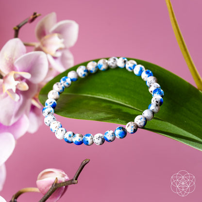 The Rare K2 Stone Bracelet of Spiritual Adventure | 6mm by Conscious Items, with white, blue, and black speckled beads, is displayed on a green leaf with orchid flowers and a pink background.