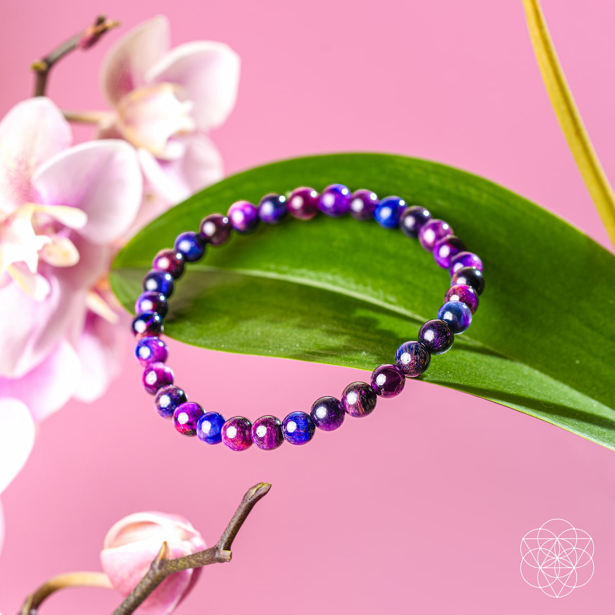 The Destiny Activator - All-Galaxy Tiger’s Eye Bracelet | 6mm by Conscious Items, featuring purple, blue, and pink stones, rests on a green leaf with pink orchid flowers in the background.