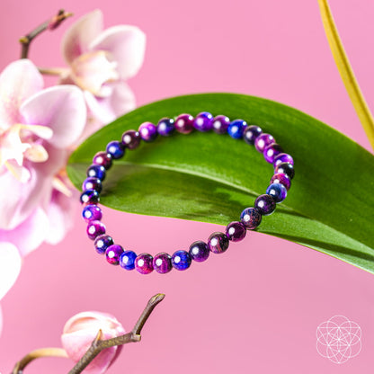 The Destiny Activator - All-Galaxy Tiger’s Eye Bracelet | 6mm by Conscious Items, featuring purple, blue, and pink stones, rests on a green leaf with pink orchid flowers in the background.