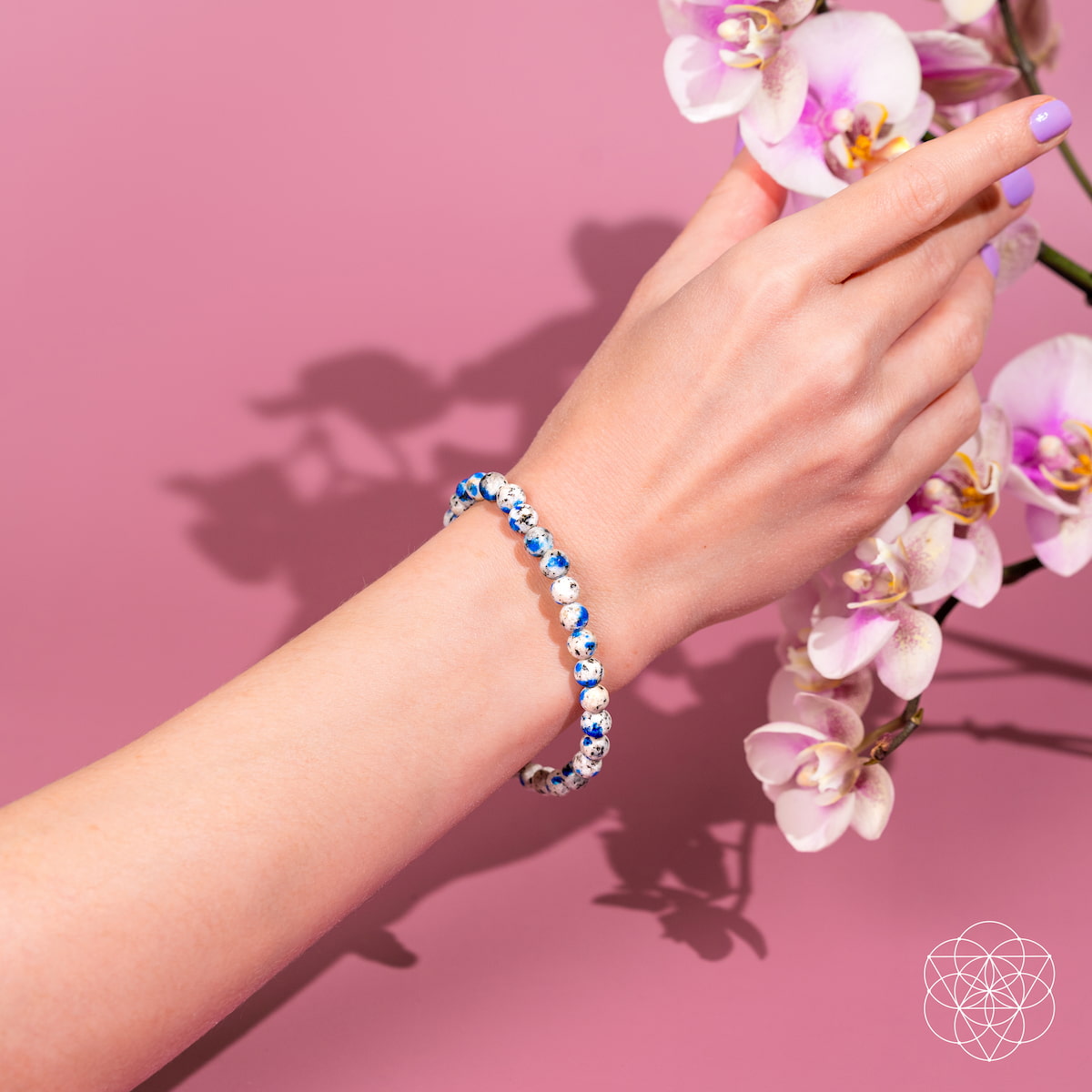 A hand adorned with the Conscious Items Rare K2 Stone Bracelet of Spiritual Adventure | 6mm reaches toward light pink orchids, set against a solid pink background.