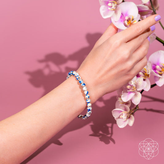 A hand adorned with the Conscious Items Rare K2 Stone Bracelet of Spiritual Adventure | 6mm reaches toward light pink orchids, set against a solid pink background.