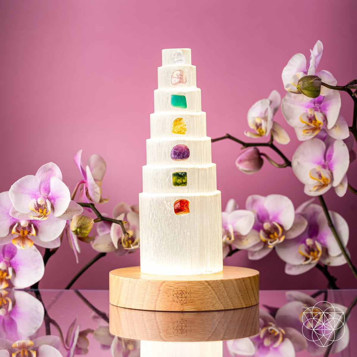 The Stress Shield Lamp - 7 Stones of Healing by Conscious Items features a tiered selenite tower with rare healing crystals on each level atop a wooden base, surrounded by pink and white orchids, set against a pink background.