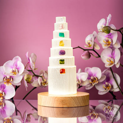 The Stress Shield Lamp - 7 Stones of Healing by Conscious Items features a tiered selenite tower with rare healing crystals on each level atop a wooden base, surrounded by pink and white orchids, set against a pink background.