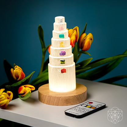 The Stress Shield Lamp - 7 Stones of Healing by Conscious Items sits on a wooden base next to yellow tulips on a white surface, with its remote control in the foreground and a blue background.