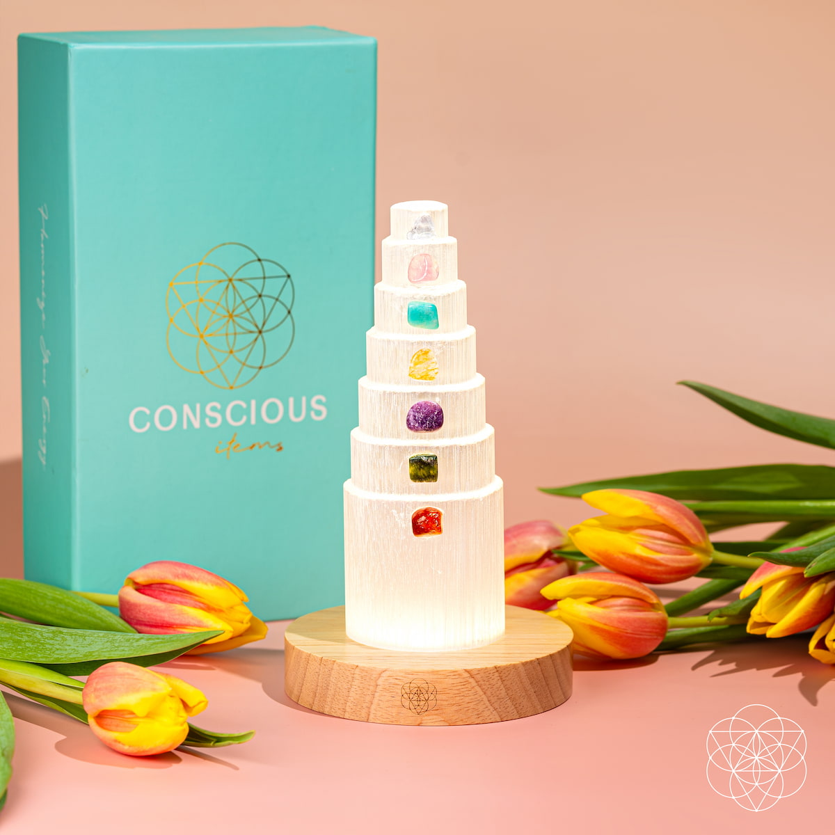 The Stress Shield Lamp - 7 Stones of Healing by Conscious Items features a tiered selenite crystal tower with rare crystals on each level, set on a wooden base and surrounded by orange tulips. A turquoise Conscious Items box stands behind it against a peach background.