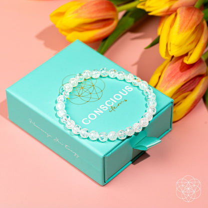 The Emotional Ice Bath – The Ice Quartz Bracelet | 6mm by Conscious Items sits on a turquoise box beside yellow and orange tulips, all set against a light pink background.