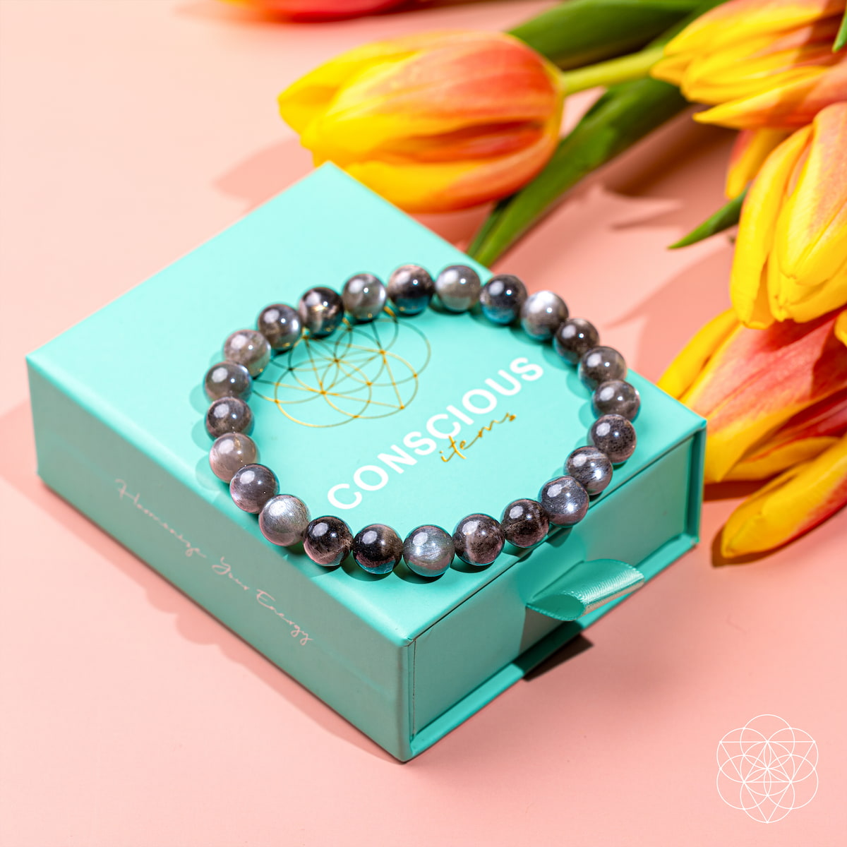The Empath’s Cord Cutter – Black Moonstone Bracelet from Conscious Items rests on a turquoise CONSCIOUS box, ideal for celebrating new beginnings. Yellow and orange tulips are nearby on a pale pink surface.