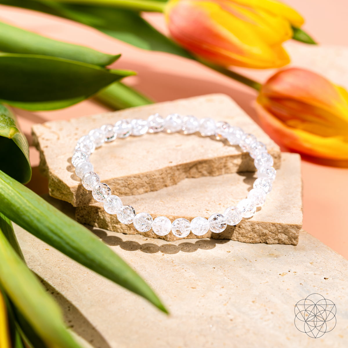 The Emotional Ice Bath – The Ice Quartz Bracelet | 6mm by Conscious Items, a reset stone for emotional clarity, is displayed on beige stones among green leaves and orange-yellow tulips, with a geometric logo on a soft peach background.