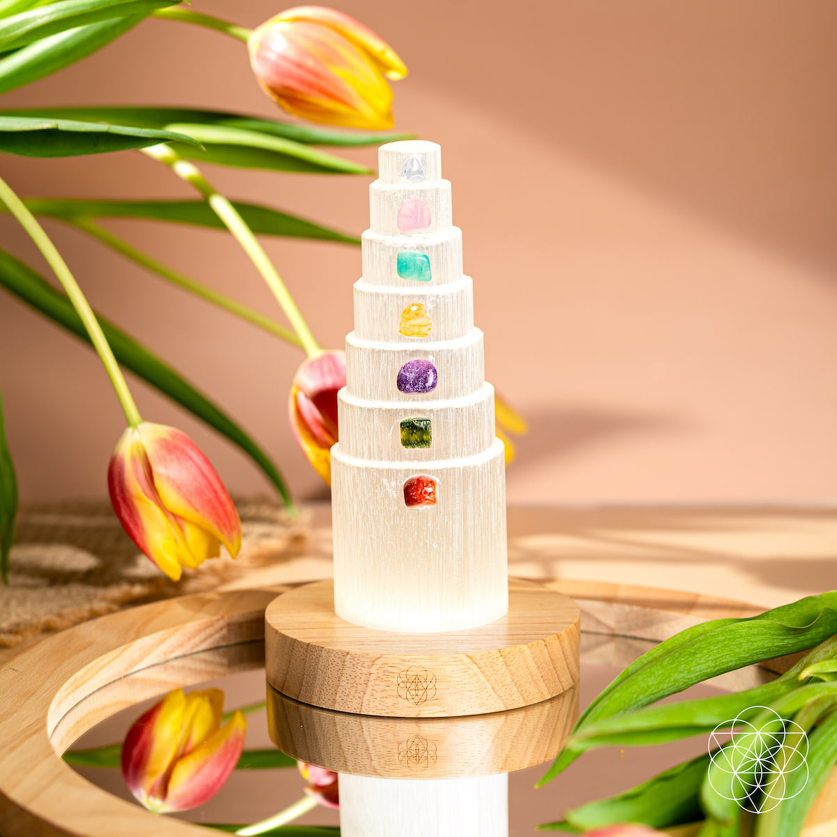 The Stress Shield Lamp - 7 Stones of Healing by Conscious Items features a selenite tower and chakra crystals on a wooden base, accented by red and yellow tulips with green leaves, beautifully reflected on a mirrored surface.