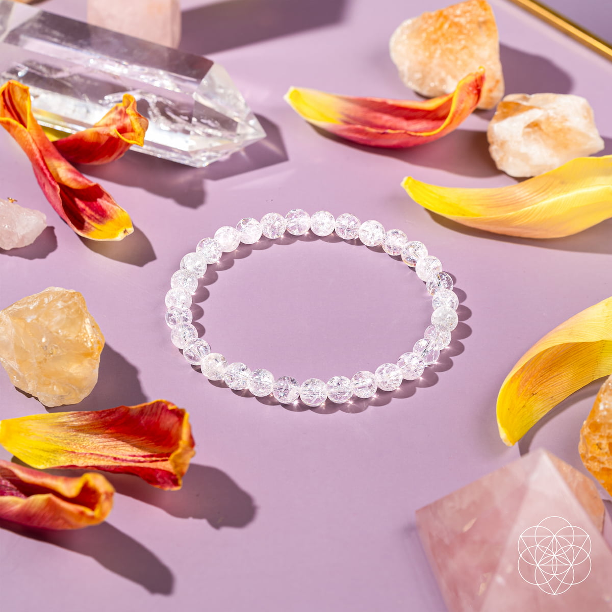 The Emotional Ice Bath – The Ice Quartz Bracelet | 6mm by Conscious Items is displayed on lavender, surrounded by colorful petals, quartz crystals, reset stones, and orange minerals. A geometric white logo is in the lower right corner.