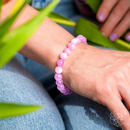 A person wearing Conscious Items' The Heart Reset - Rare Pink Kunzite Bracelet sits among green leaves, featuring lavender-painted nails and light blue jeans. Pink Kunzite is known as a heart opener that supports emotional healing.