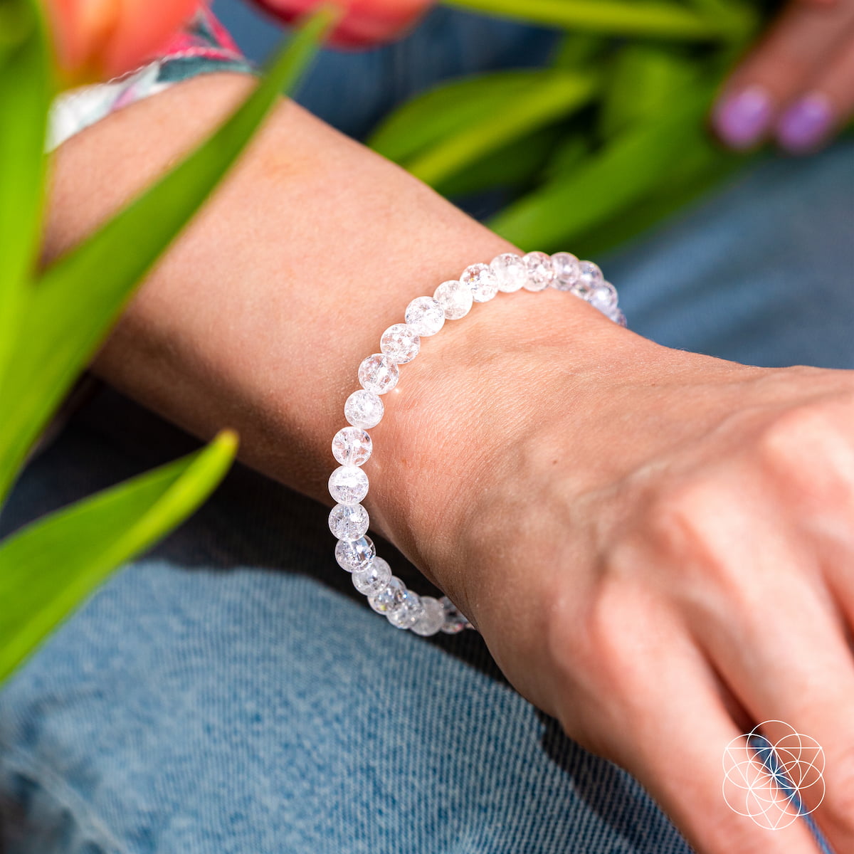 A wrist adorned with the Emotional Ice Bath – The Ice Quartz Bracelet | 6mm by Conscious Items rests among green leaves and orange flowers. Blue jeans and light purple nail polish complete this look of emotional clarity.