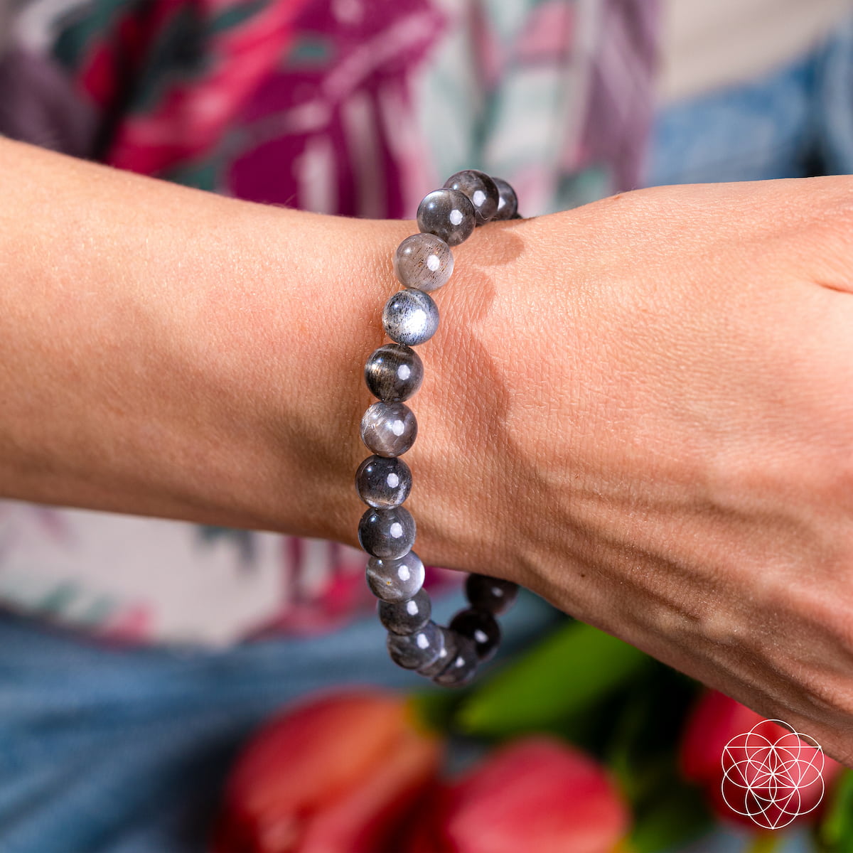 A close-up of a wrist adorned with The Empath’s Cord Cutter – Black Moonstone Bracelet by Conscious Items, showcasing shiny dark gray and light blue stones. The blurred background features colorful fabric and red tulips.