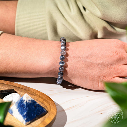 A person wears The Empath’s Cord Cutter – Black Moonstone Bracelet by Conscious Items, designed for new beginnings, while their arm rests on a light surface near a wooden dish of crystals and green leaves in the foreground.